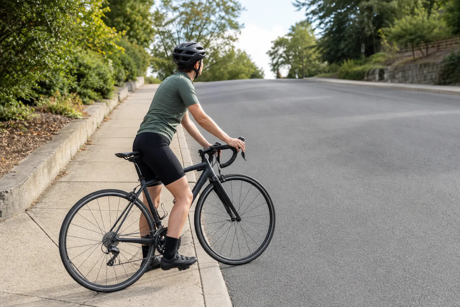 A helmeted beginner road cyclist has stopped safely near the curb on an uphill road with one foot down and both hands on the brake hoods
